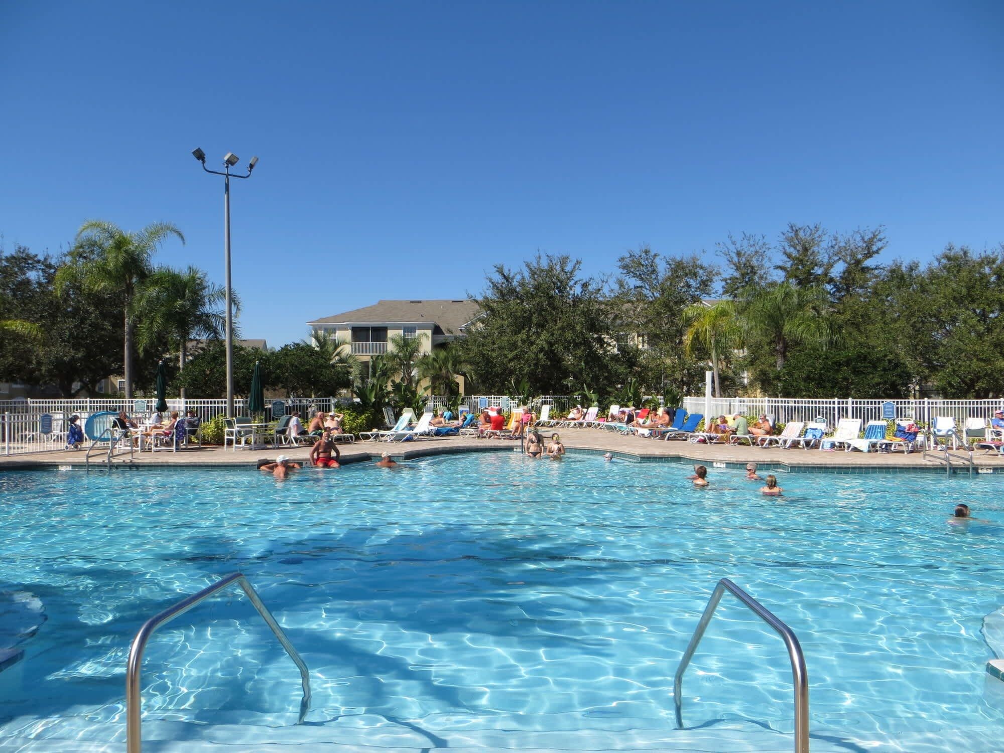 Piscina com água cristalina, rodeada por espreguiçadeiras e guarda-sóis, com árvores e um edifício ao fundo.