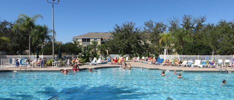 A swimming pool with clear water, surrounded by lounge chairs and umbrellas, with trees and a building in the background.