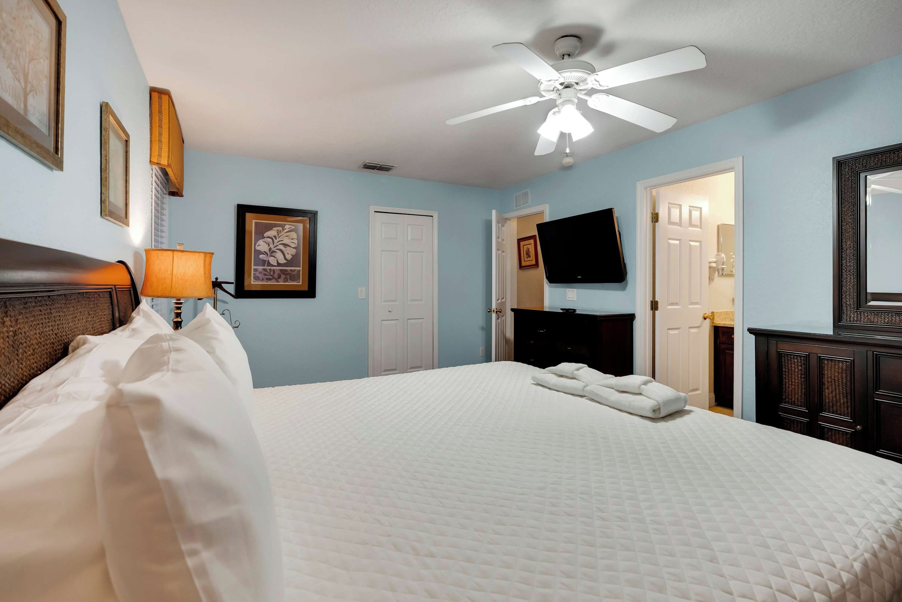 A hotel room with a bed, a ceiling fan, a television, and a door leading to a bathroom.