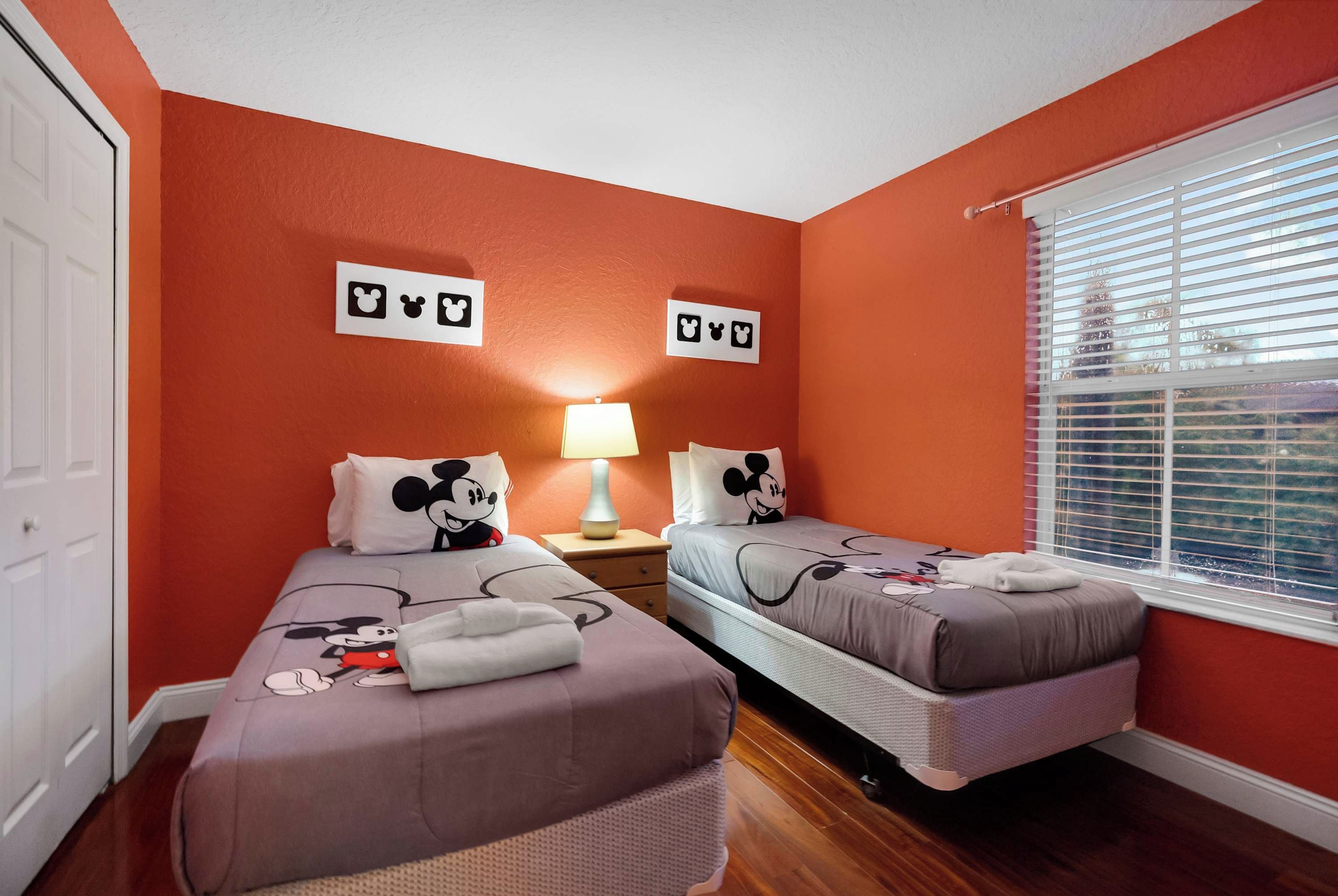 A hotel room with two beds, each with Mickey Mouse bedding, a nightstand with a lamp, and a window with blinds.