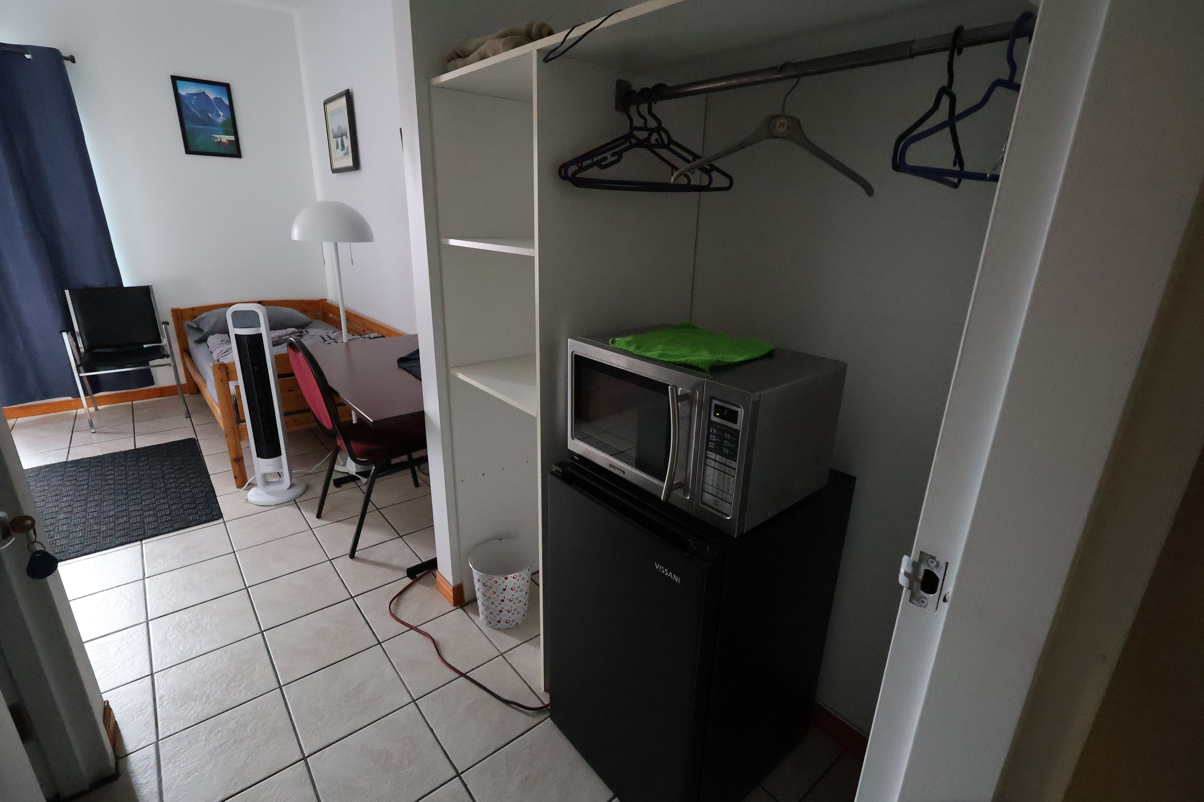 Une petite pièce comprenant un lit, un bureau, une chaise, une lampe, un four à micro-ondes et une petite poubelle.