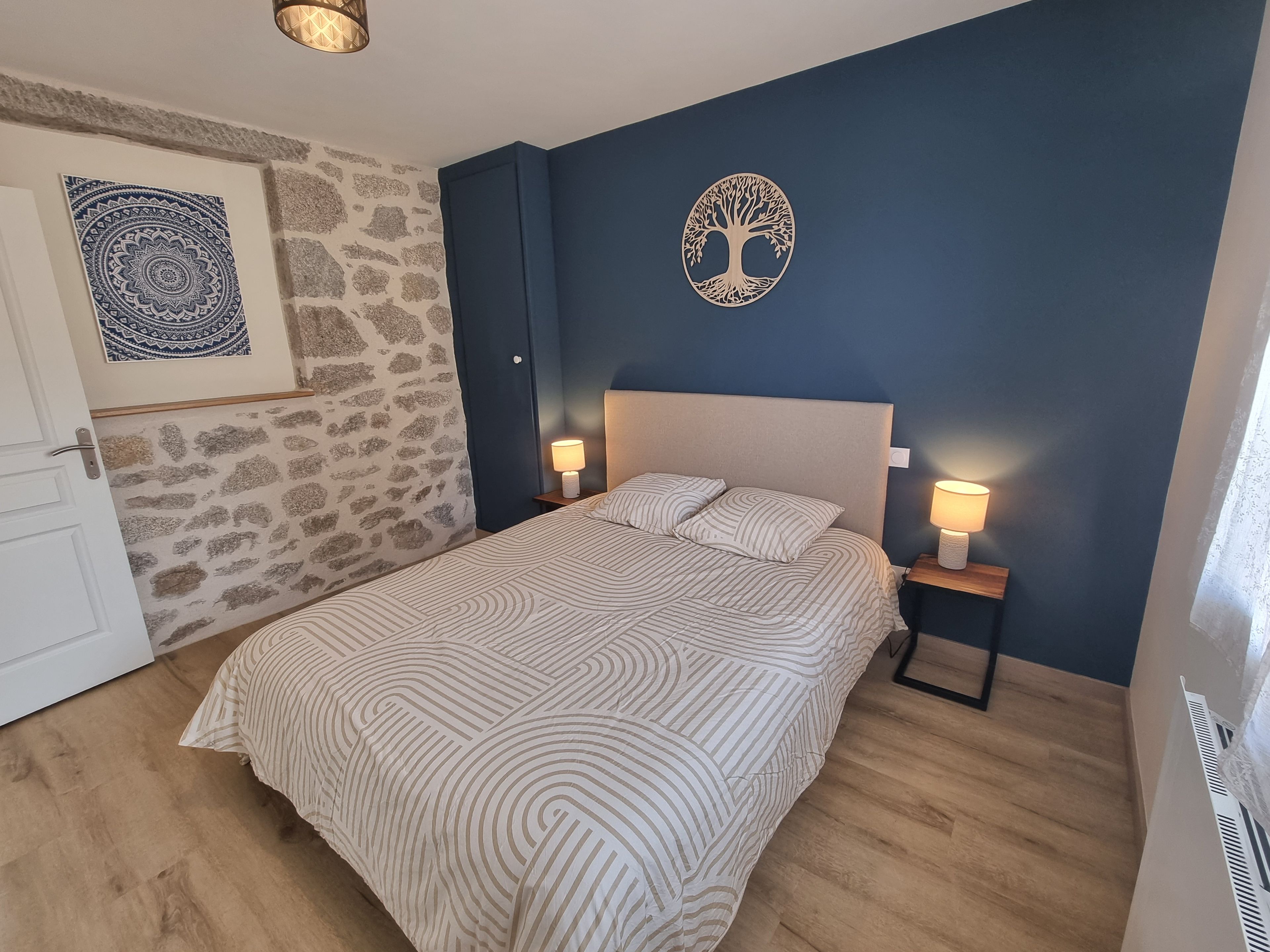 Quarto com cama, duas luminárias de cabeceira, tapeçaria na parede e um recorte decorativo de árvore.