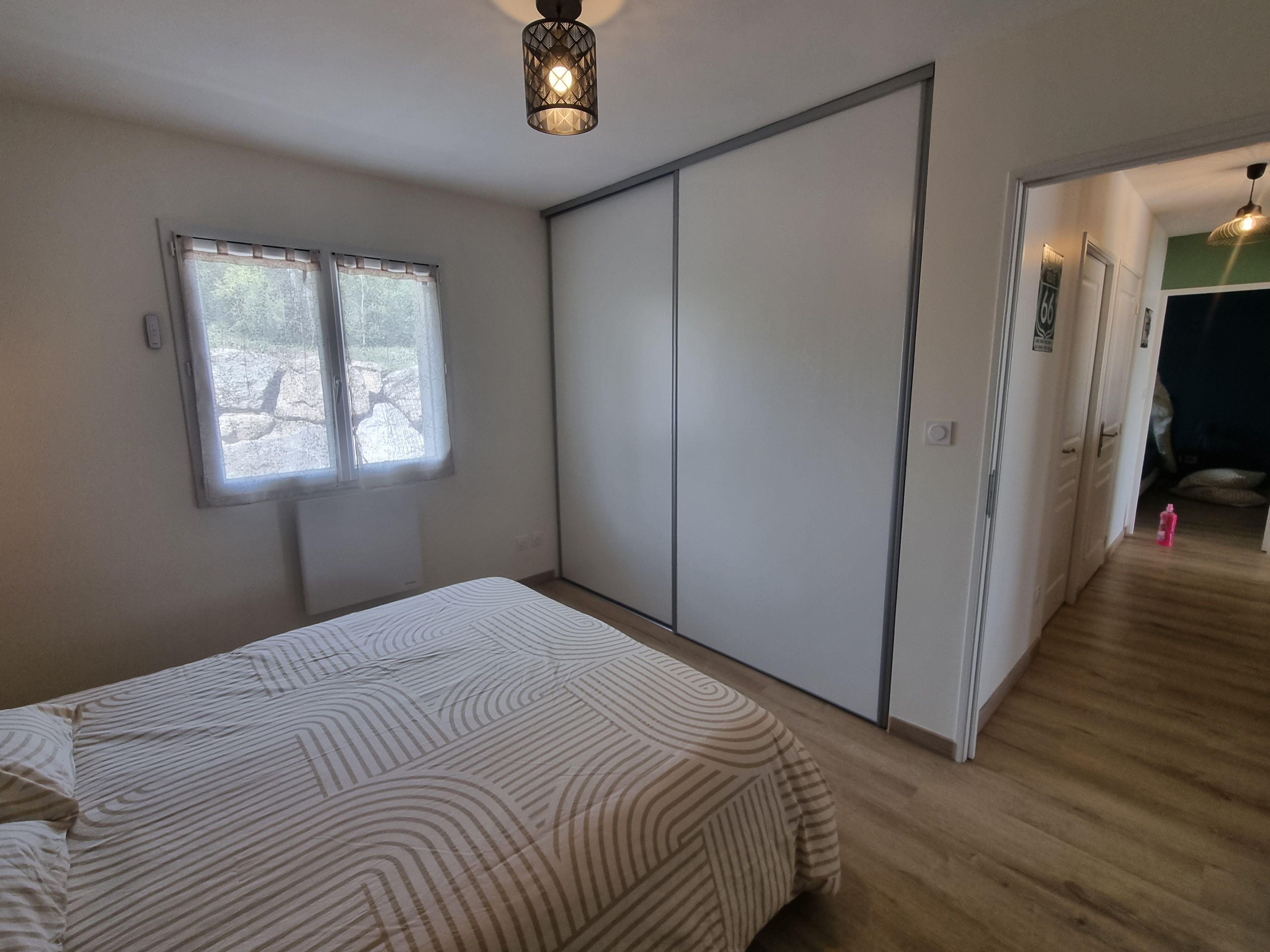 A bedroom with a bed, a window, a wardrobe, and a door leading to another room.