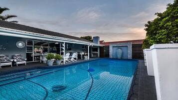 A swimming pool with a tiled deck, lounge chairs, and a covered seating area with tables and chairs.