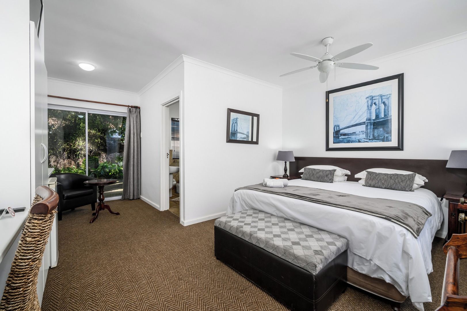 Quarto de hotel com cama, escrivaninha, cadeira, ventoinha no teto e janela com vista para uma ponte.
