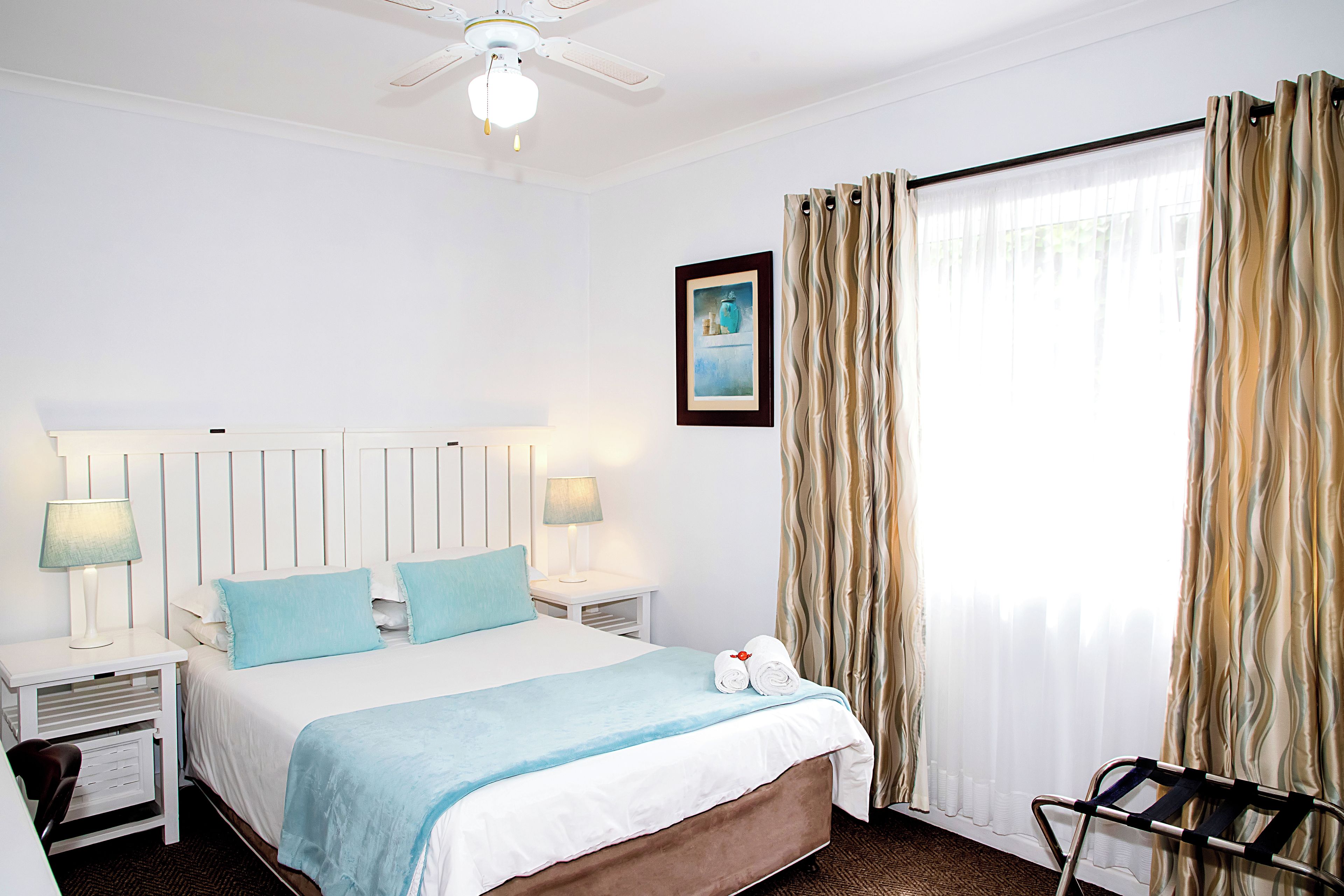 Habitación de hotel con cama, dos lámparas de noche, ventilador de techo, ventana con cortinas y un cuadro en la pared.