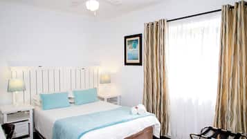 Habitación de hotel con cama, dos lámparas de noche, ventilador de techo, ventana con cortinas y un cuadro en la pared.