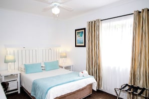 Habitación de hotel con cama, dos lámparas de noche, ventilador de techo, ventana con cortinas y un cuadro en la pared.