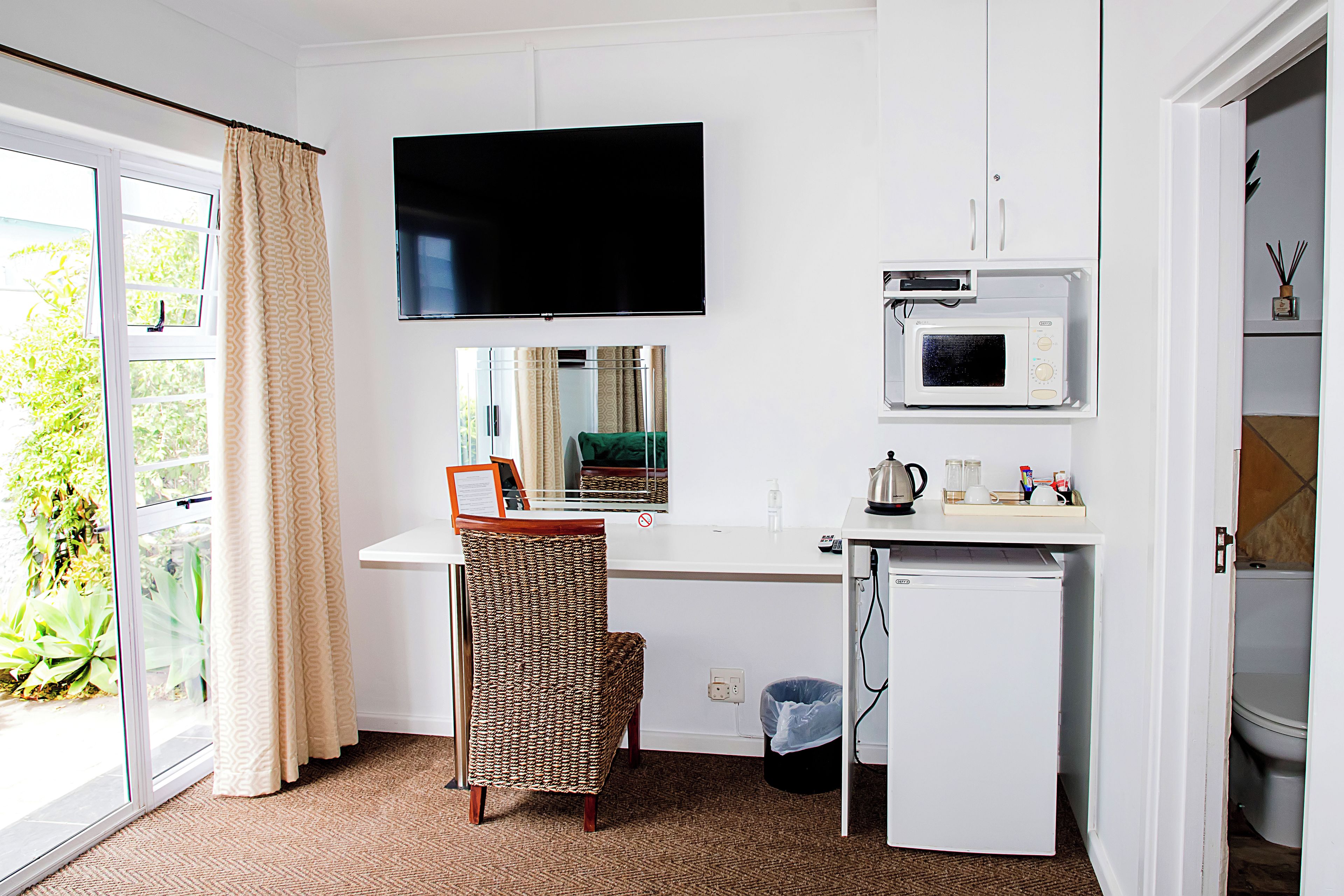 Een compacte kamer met een flatscreen televisie boven een bureau, een magnetron, een kleine koelkast, en een rieten stoel.