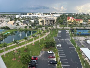 A parking lot with several cars, a tennis court, and a body of water with palm trees.