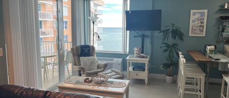 A living room with a leather sofa, a dining table with chairs, a TV, and a view of the ocean.