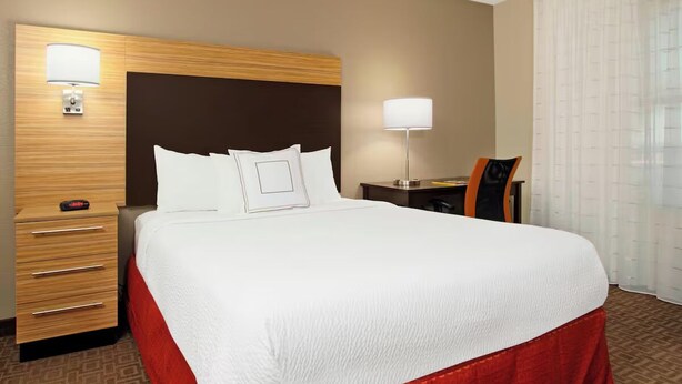 Une chambre d’hôtel avec un grand lit, une table de chevet avec une lampe, un bureau avec une chaise et une fenêtre avec des rideaux.