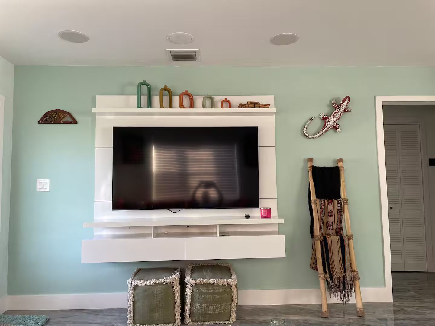 Un salon avec un téléviseur à écran plat fixé sur un meuble de rangement blanc, une décoration murale et un fauteuil traditionnel en osier.