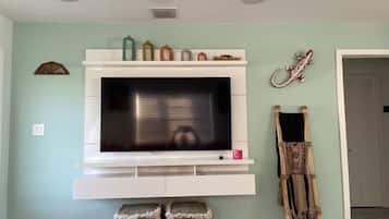 A living room with a flat-screen TV mounted on a white entertainment unit, a decorative wall hanging, and a traditional woven chair.