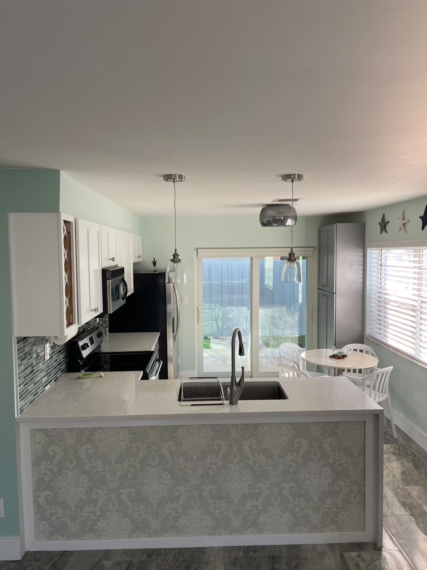 A modern kitchen with a central island, a dining area with a round table, and a view through a glass door.