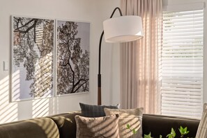 Un salon comprenant un canapé, deux œuvres d’art représentant des arbres encadrées, une lampe sur pied et une fenêtre avec des stores.