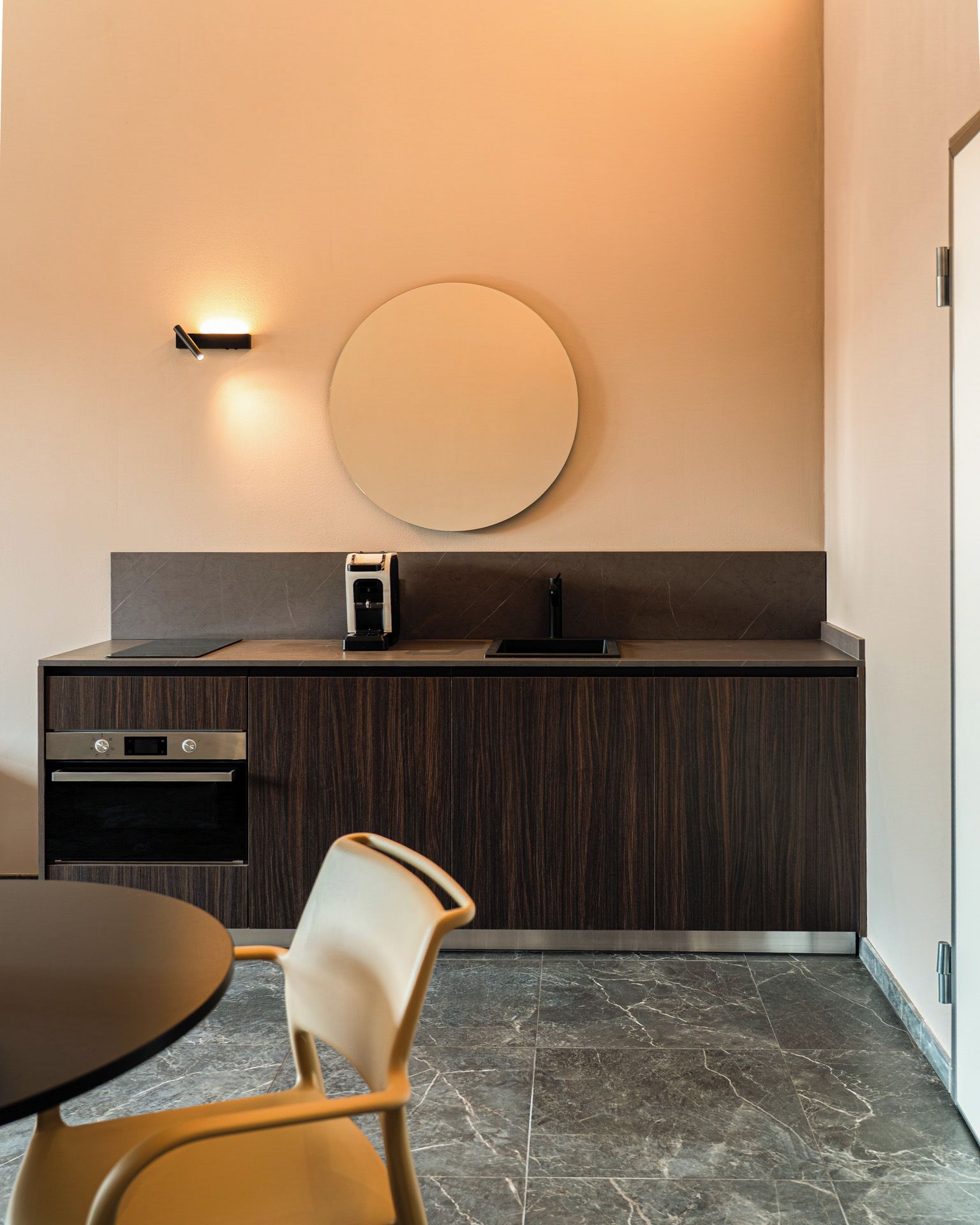 Une cuisine moderne avec un grand miroir rond, des meubles de couleur bois foncé, un appareil à café intégré et une table ronde accompagnée d’une chaise.
