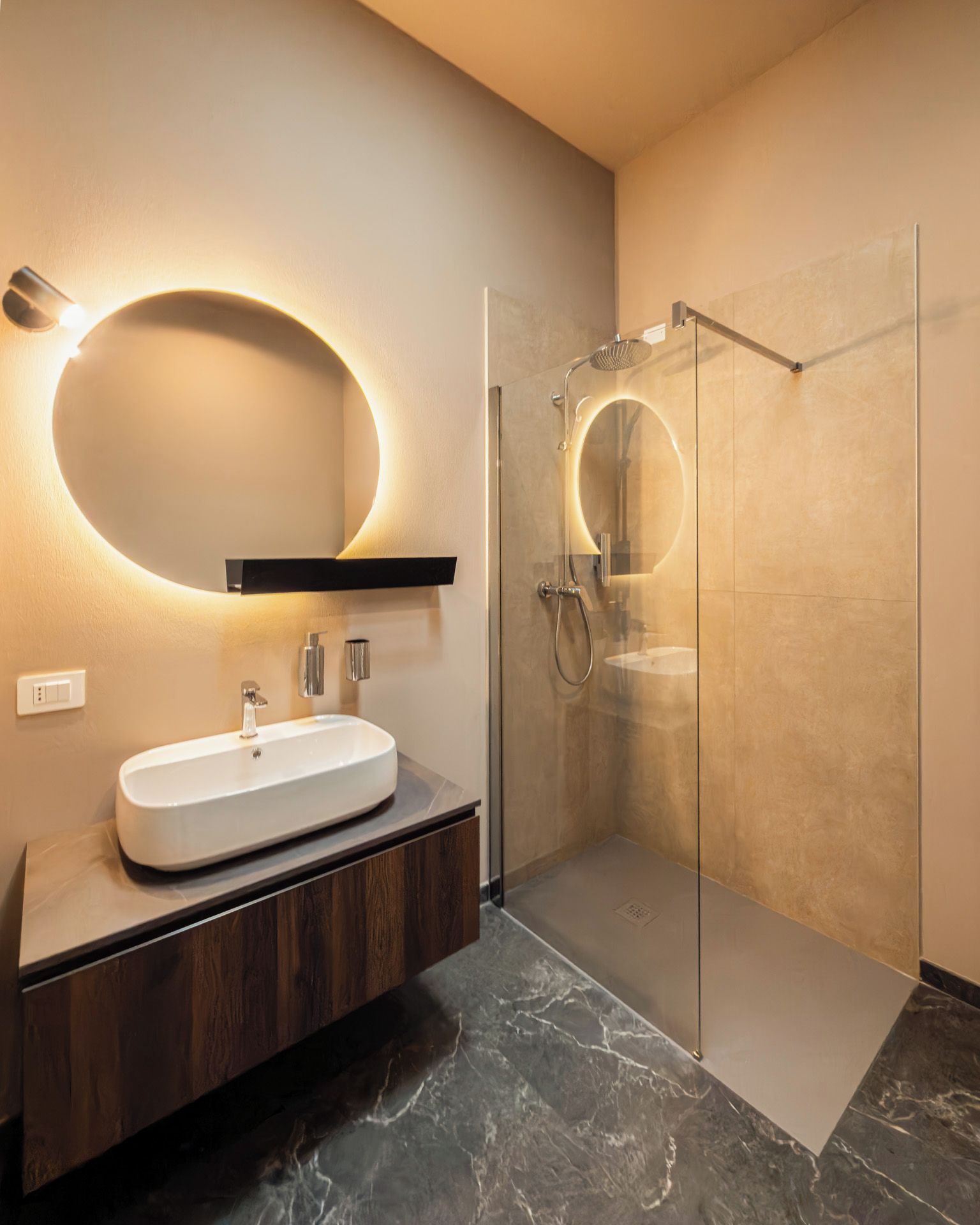 Ein modernes Badezimmer mit einer ein Glasfach raumseitig geschlossenen Dusche, einem weißen Waschbecken und einem wandmontierten Spiegel mit runder Beleuchtung.