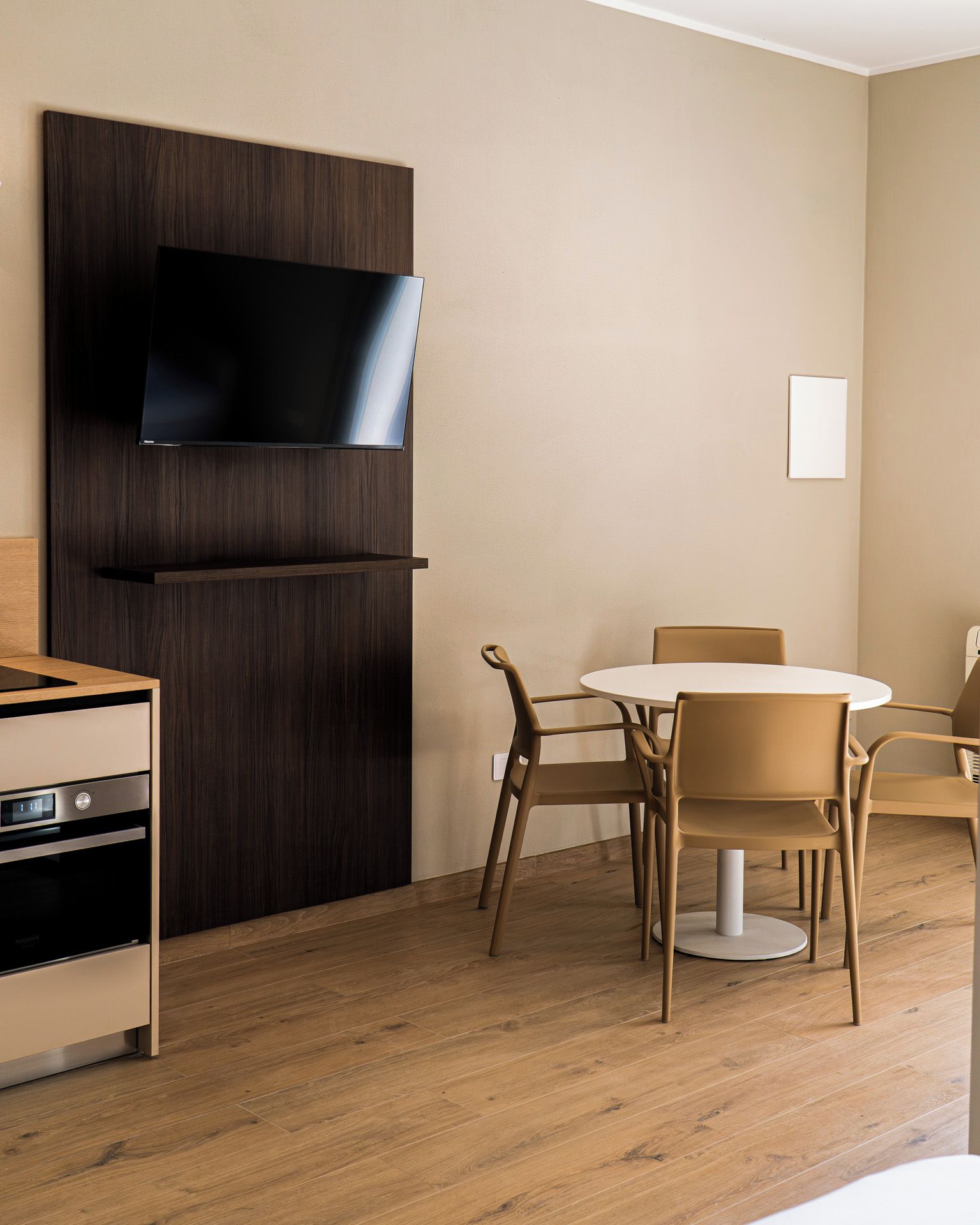 Une chambre d’hôtel dotée d’un téléviseur à écran plat fixé sur un panneau en bois foncé, d’une table de salle à manger ronde avec quatre chaises, et d’une kitchenette équipée d’une cuisinière et d’un four.