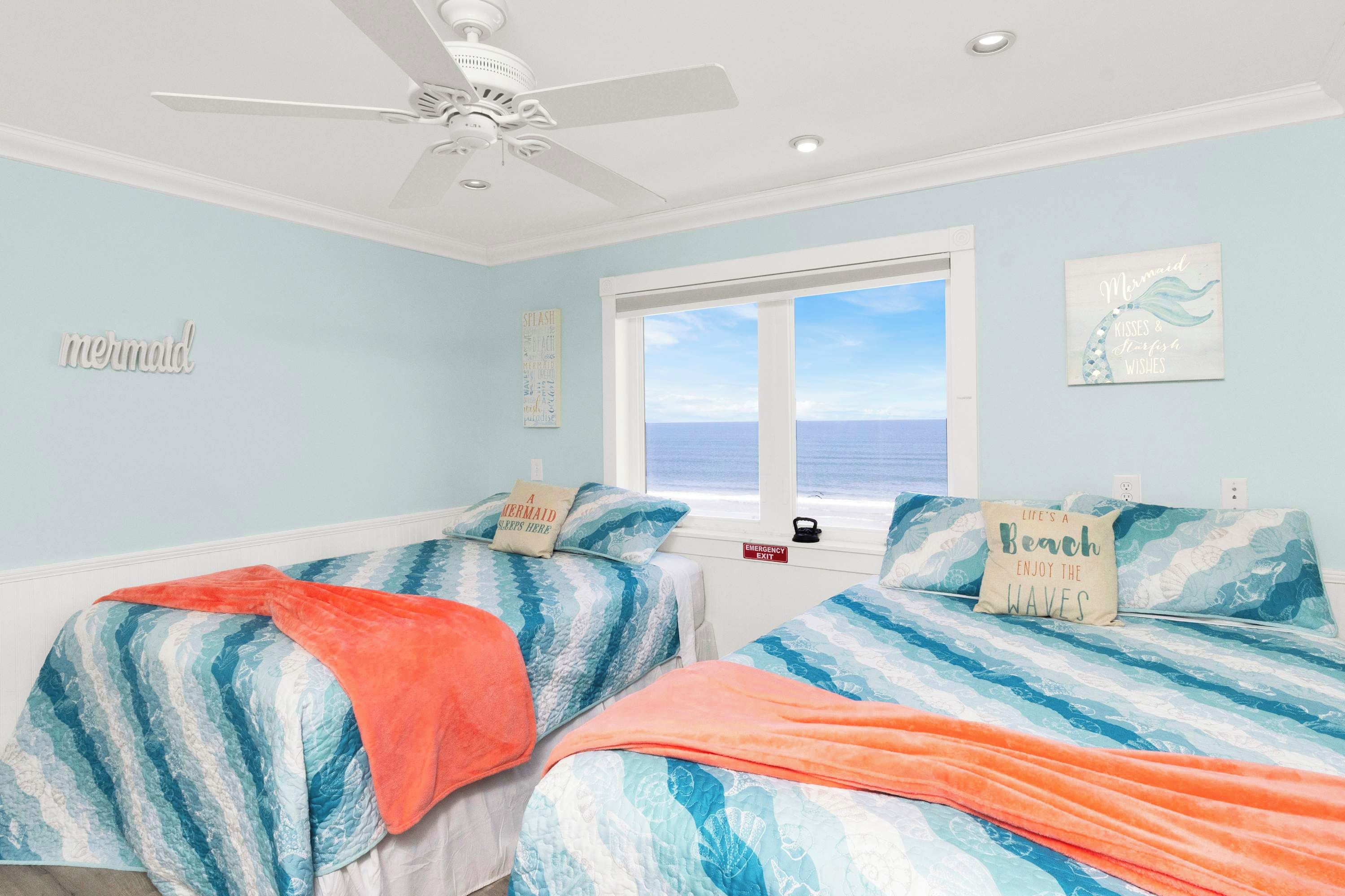 Une chambre avec deux lits, un ventilateur de plafond, une fenêtre donnant sur la mer et un panneau indiquant la sortie de secours.