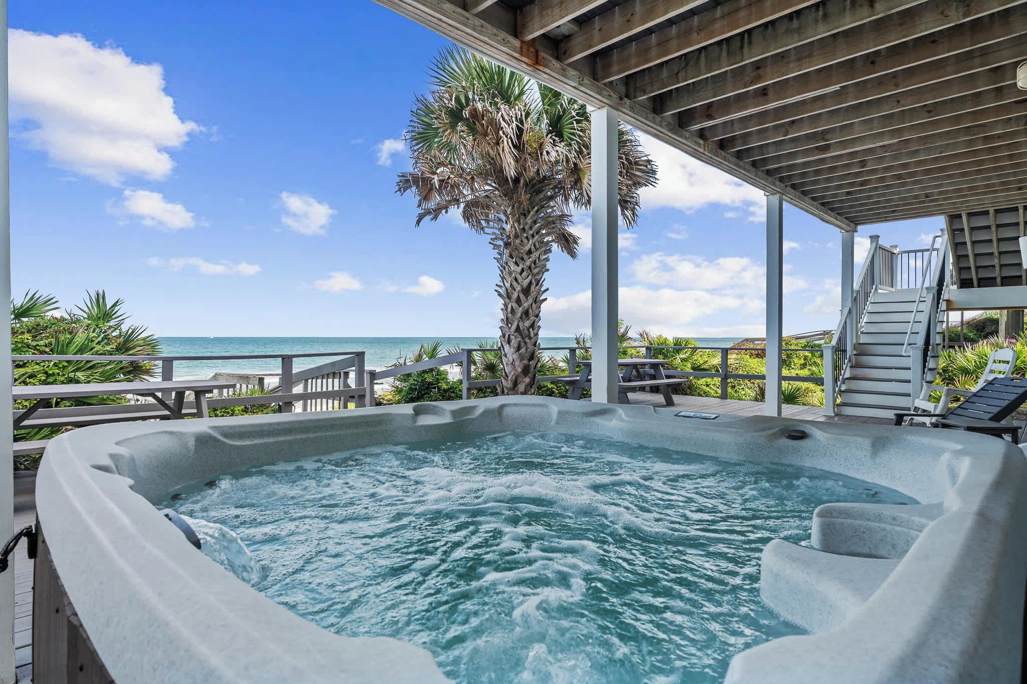 A hot tub with a view of the ocean, surrounded by a wooden deck and lounge chairs.