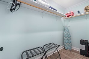 Quarto com cama, varal de roupa, mala e prateleira com diversos objetos.