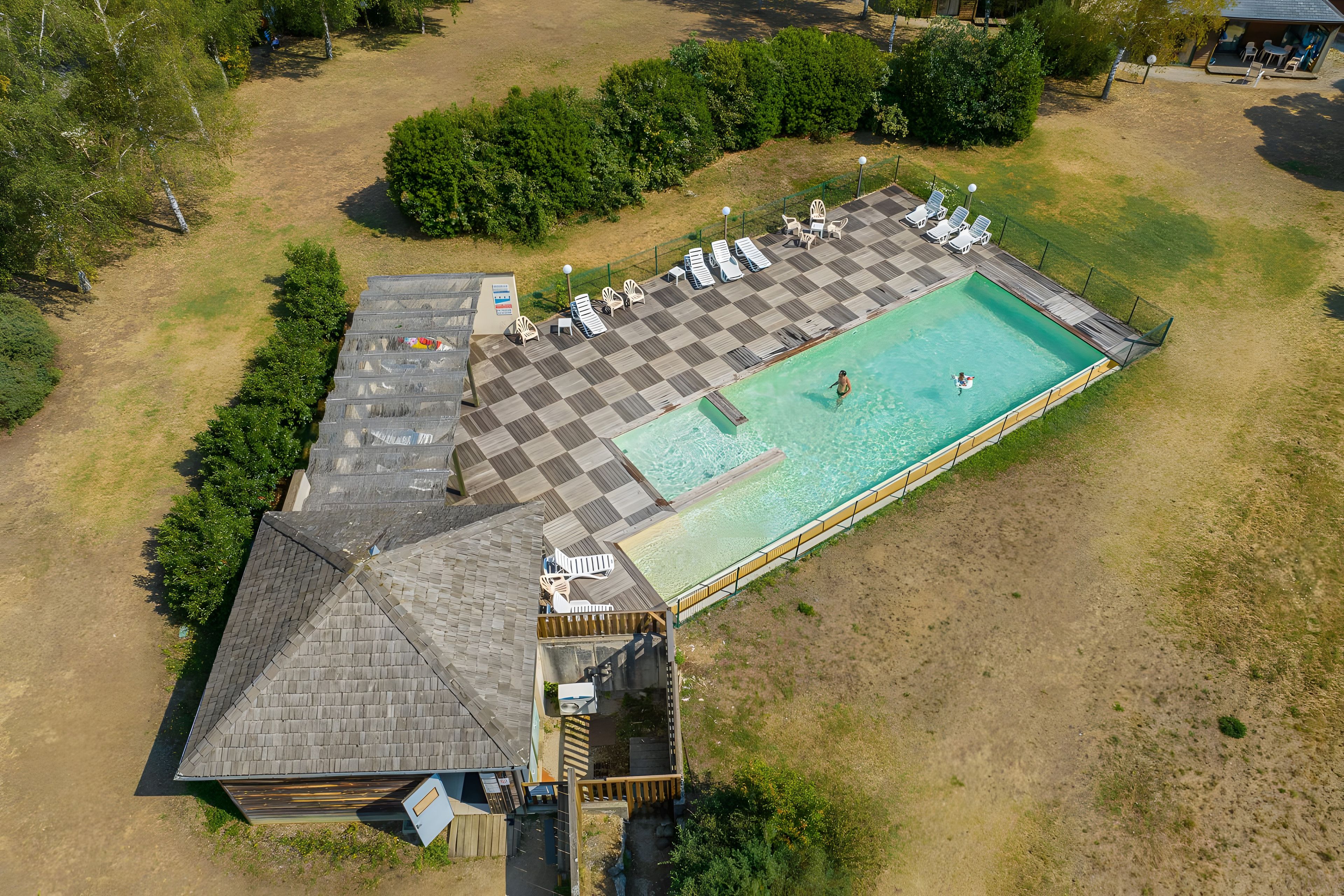 Eine Vogelperspektive auf einen Poolbereich mit Schachbrettmuster-Terrasse, Liegestühlen und einem kleinen Gebäude.