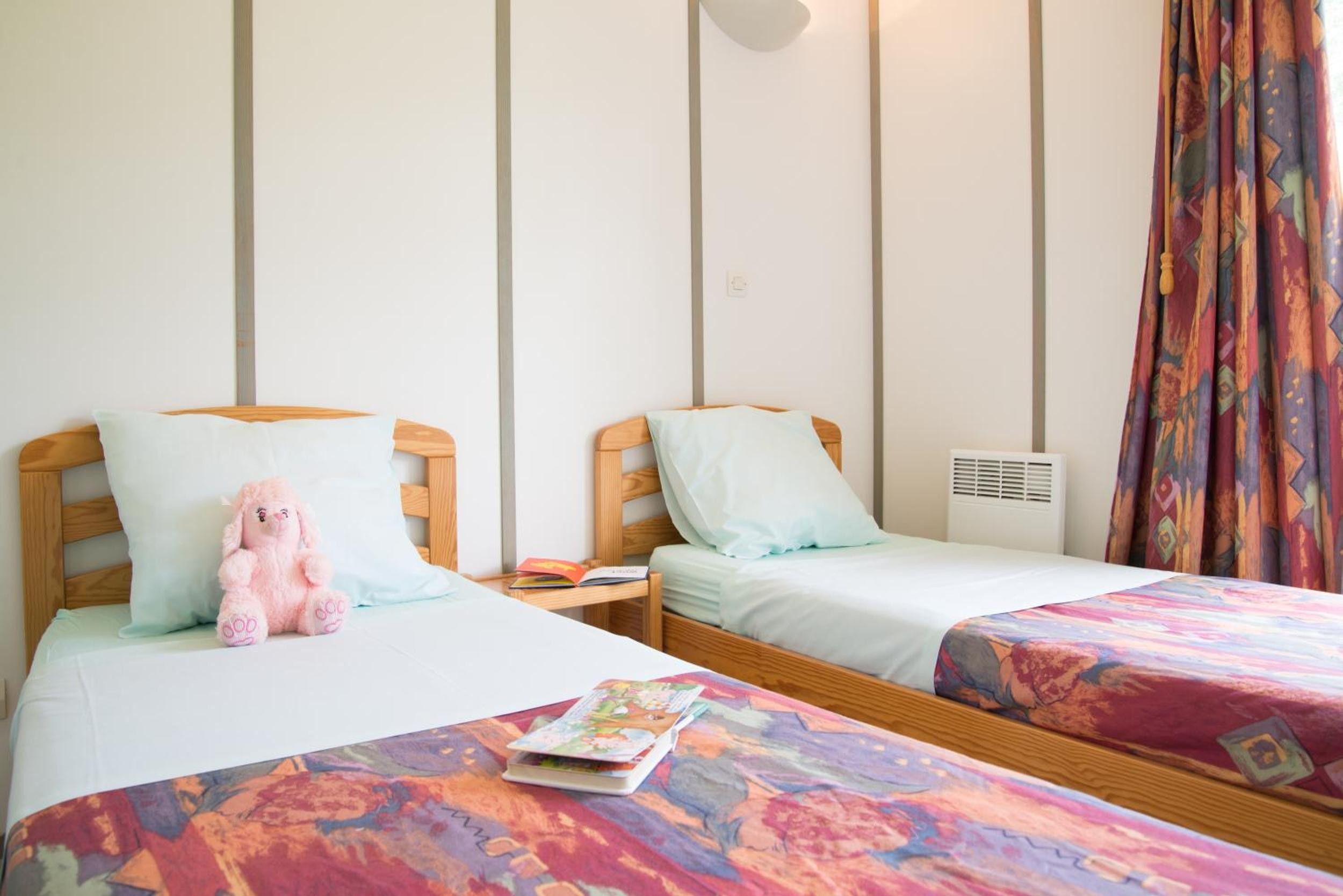 Une chambre d’hôtel avec deux lits, chacun doté d’une tête de lit en bois, d’une petite table de chevet et d’une table de nuit. La chambre comprend une fenêtre avec des rideaux, un climatiseur fixé au mur et une couvre-lit aux couleurs vives.