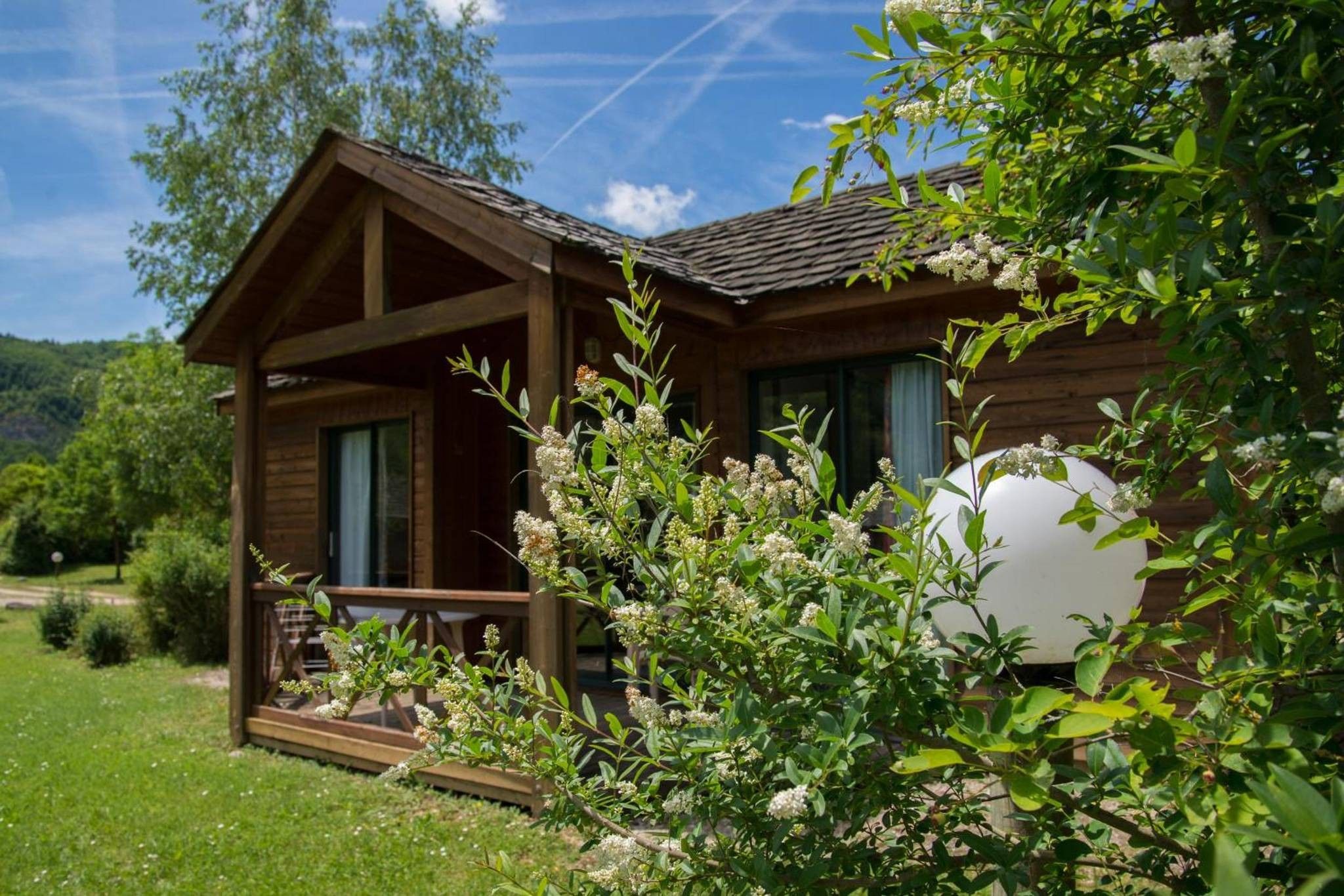 A wooden cabin with a porch, surrounded by greenery and a clear sky.
