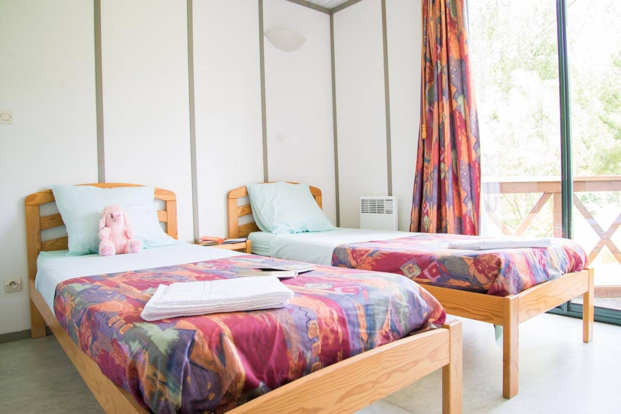 Une chambre avec deux lits en bois, chacun recouvert d’une couverture colorée et agrémenté d’un oreiller. Il y a une fenêtre avec un rideau, ainsi qu’une petite table sur laquelle se trouvent un livre et un jouet en peluche.