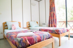 Une chambre avec deux lits en bois, chacun recouvert d’une couverture colorée et agrémenté d’un oreiller. Il y a une fenêtre avec un rideau, ainsi qu’une petite table sur laquelle se trouvent un livre et un jouet en peluche.