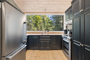 A modern kitchen with dark cabinetry, a stainless steel refrigerator, a built-in oven, and a window providing a view of trees. - Spa & Nature Escape | L'Ame des Bois (Grenville-sur-la-Rouge)
