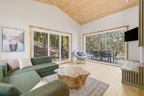 A modern living room with a green sofa, a round coffee table, and a large window overlooking a forest. - Spa & Nature Escape | L'Ame des Bois (Grenville-sur-la-Rouge)