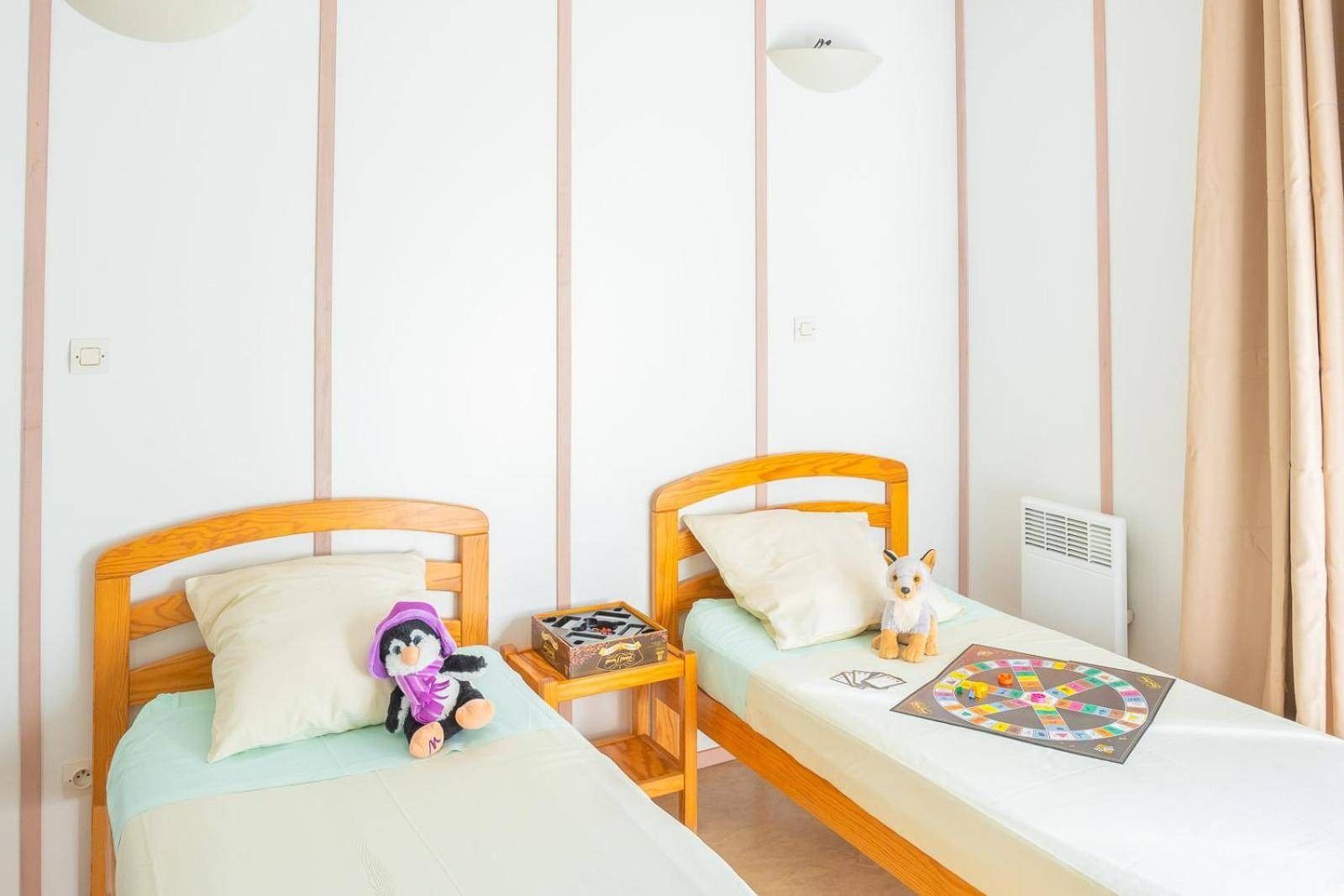 Une chambre avec deux lits simples, une petite table sur laquelle est posé un jeu de société et un ours en peluche sur l’un des lits.