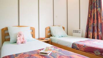 Deux lits simples avec des cadres en bois et une literie blanche, un ours en peluche rose sur le lit de gauche et une couvre-lit colorée sur le lit de droite.