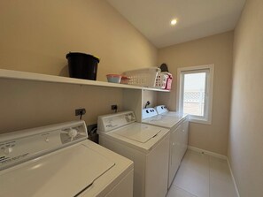 A laundry room with two front-loading washers and dryers, a shelf with laundry baskets, and a window with blinds. - Three Harbour Villa in Samaan Grove (Canaan, Tobago)