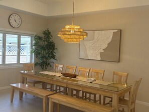 A dining room with a glass-top table, wooden chairs, a bench, a hanging light fixture, a clock, and a large abstract painting. - Three Harbour Villa in Samaan Grove (Canaan, Tobago)