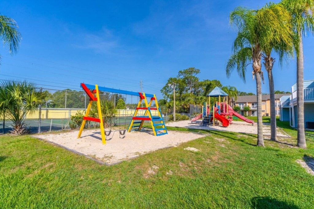 Un parque infantil con columpios y tobogán, rodeado de césped y palmeras.