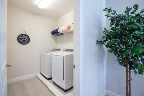 A laundry room with two white washing machines, a wall-mounted shelf with a hand holding a yellow object, and a potted plant. - 6 BD/ 6.5 BA/ Encore Resort at Reunion (404 SS) (Kissimmee)