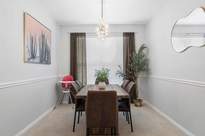 A dining room with a wooden table, chairs, a chandelier, a potted plant, and a painting on the wall.