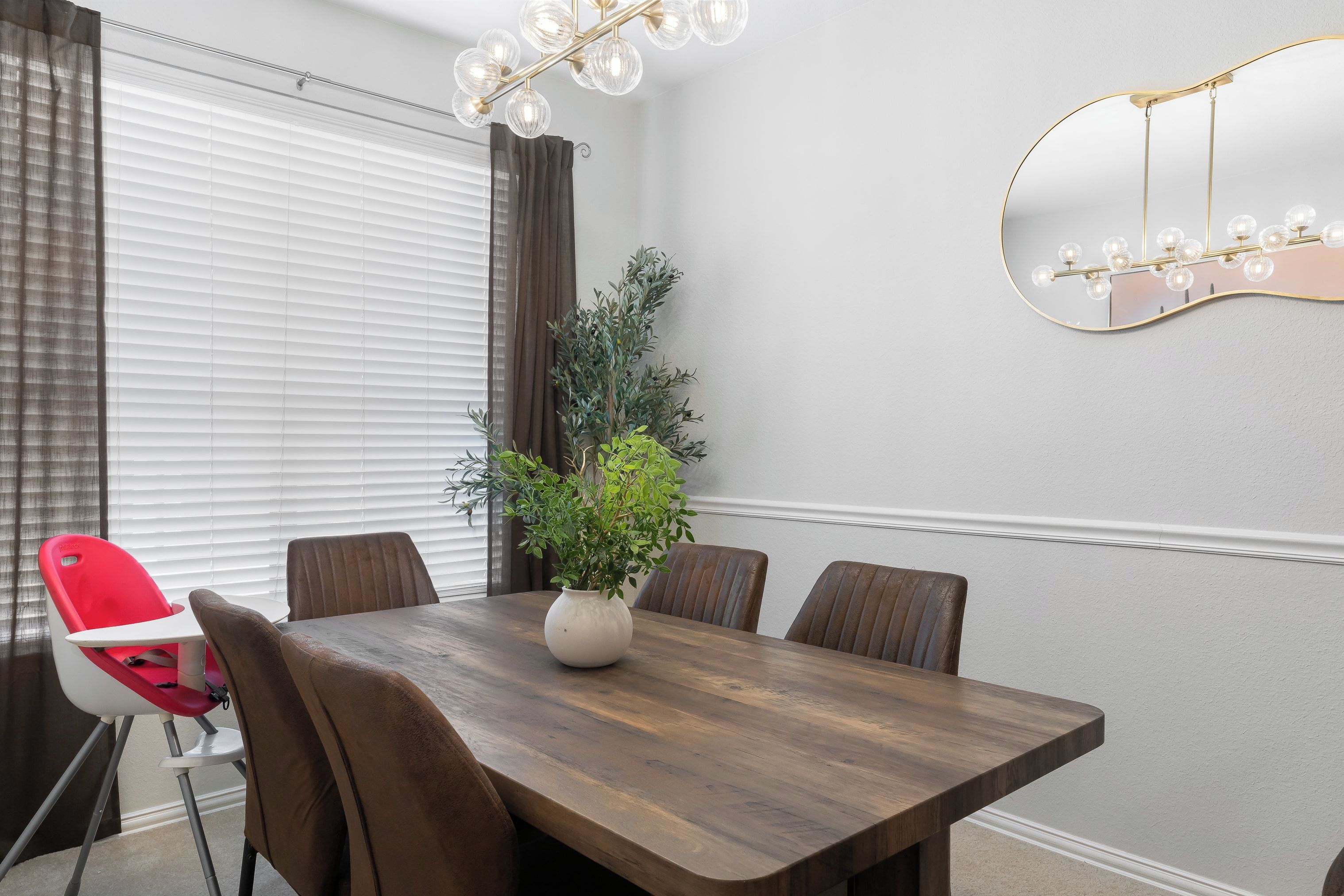 A dining area with a wooden table, six chairs, a potted plant, and a wall-mounted mirror.