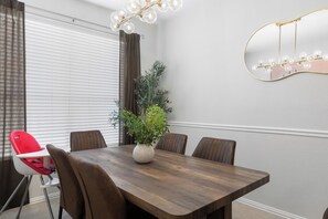 Uma sala de jantar com mesa de madeira, seis cadeiras, um vaso de planta e um espelho fixado na parede.