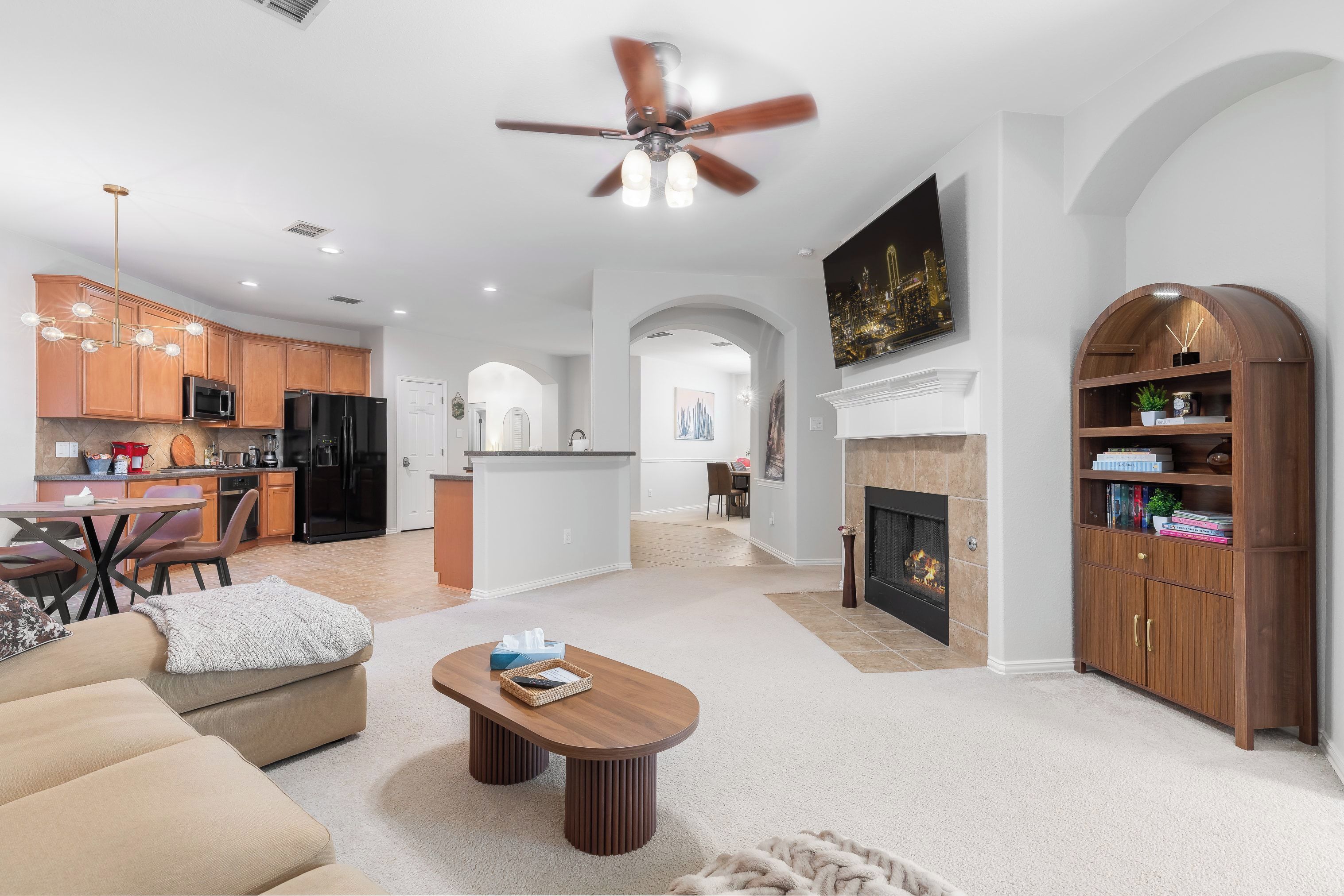 A modern living room with a fireplace, a dining area, and a kitchen with wooden cabinets and a ceiling fan.