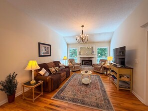 A living room with a fireplace, a chandelier, a television, and a large window. - Near Helen | Dog-Friendly Mountain Retreat + Pool (Sautee Nacoochee)