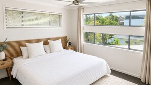 Une chambre avec un grand lit, une tête de lit en bois, une table de chevet, un radiateur fixé au mur et une fenêtre donnant sur un espace verdoyant et un immeuble.