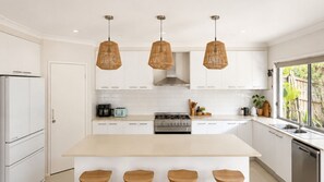 Cozinha moderna com armários brancos, uma ilha central com banquetas, fogão a gás e três luminárias de vime suspensas.