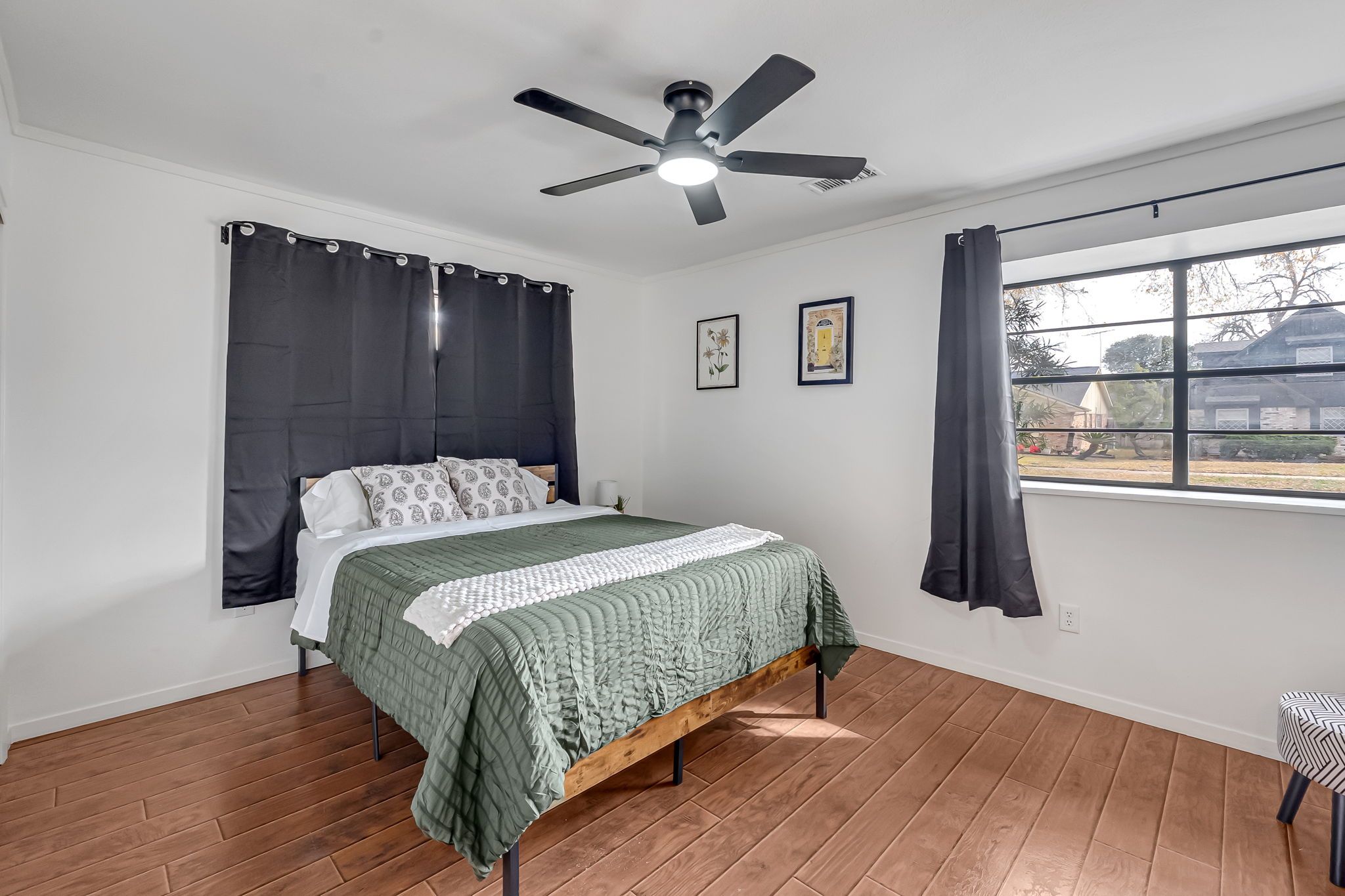 A bedroom with a bed, a ceiling fan, a window with curtains, and a view of the outside.