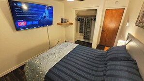 A bedroom with a bed, a TV mounted on the wall displaying a sports channel, a closet with hanging clothes, and a door leading to another room. - At Glacier's doorstep, stylish 2BR, kitchen, fire pit, BBQ, laundry, yard, wi-fi (Juneau)