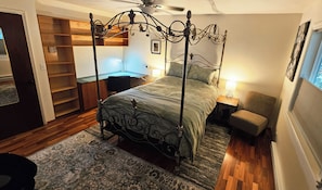 A four-poster bed with a dark metal frame, a wooden desk, a chair, and a small table with a lamp.