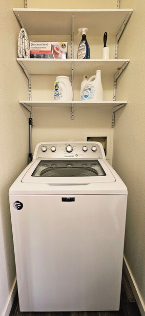 Un appareil combiné lave-linge et sèche-linge à chargement frontal, de couleur blanche, doté de boutons de commande, d’un câble d’alimentation et d’une étagère fixée au mur au-dessus, servant à ranger les produits de nettoyage.